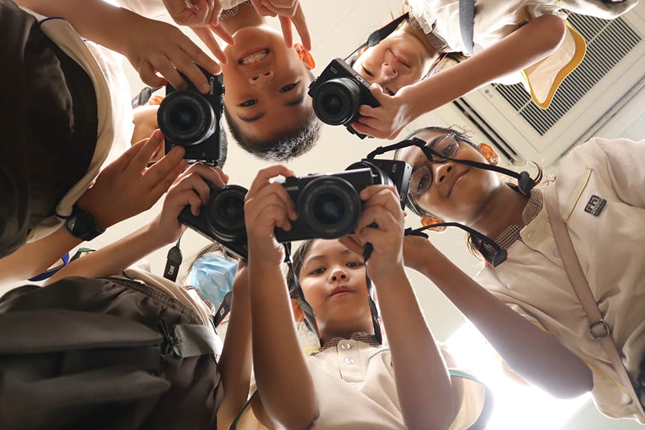 Five children in school uniforms lean into a circle, each holding a black camera, photographed from below.