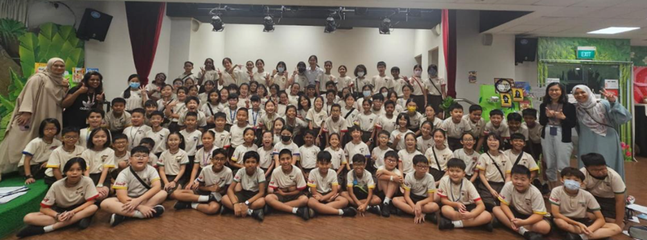 Large group of young students in matching white uniforms posing with teachers in an indoor hall.