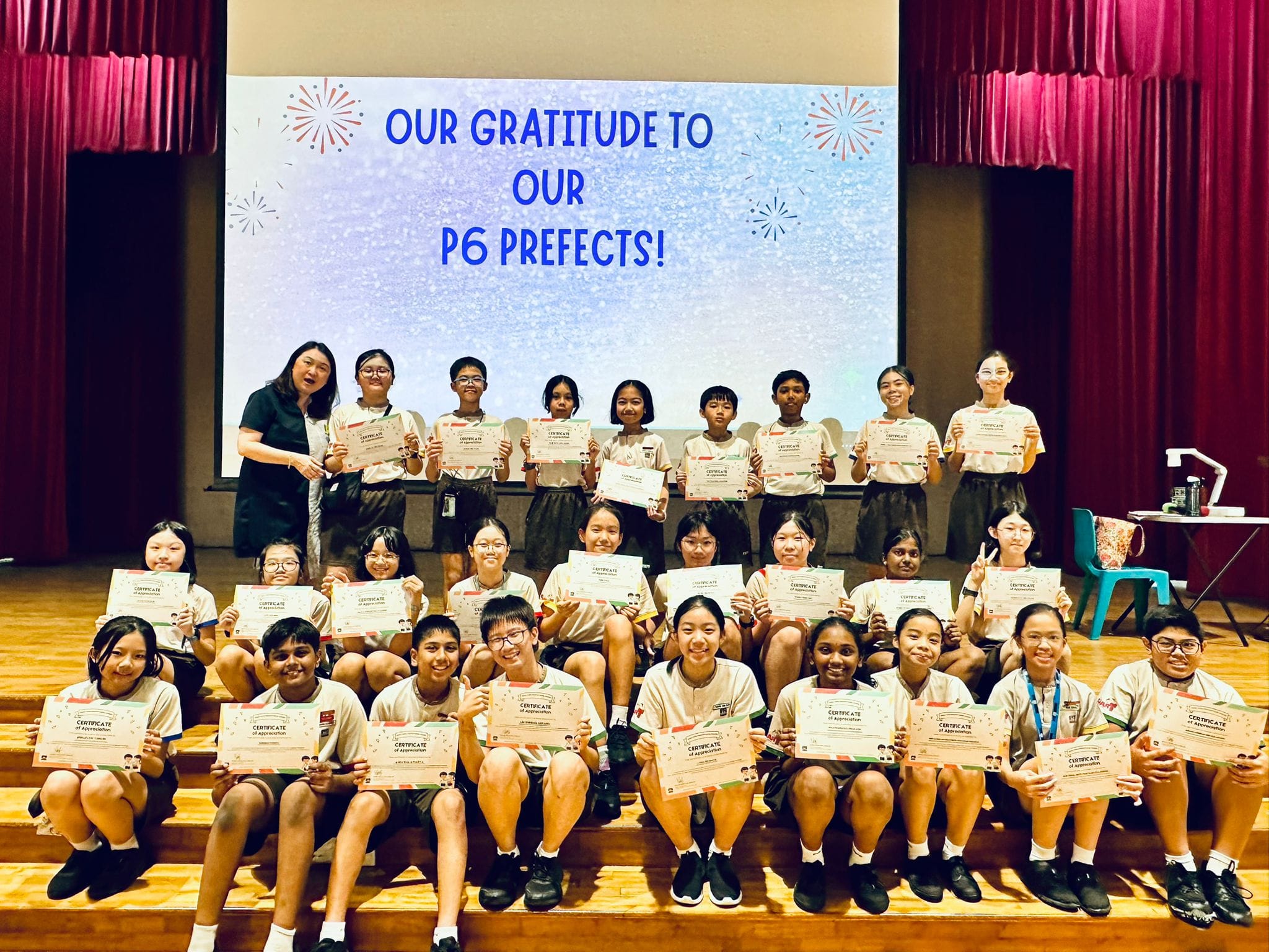 Group of P6 student prefects holding certificates on a stage, with a screen reading "Our Gratitude to Our P6 Prefects!"