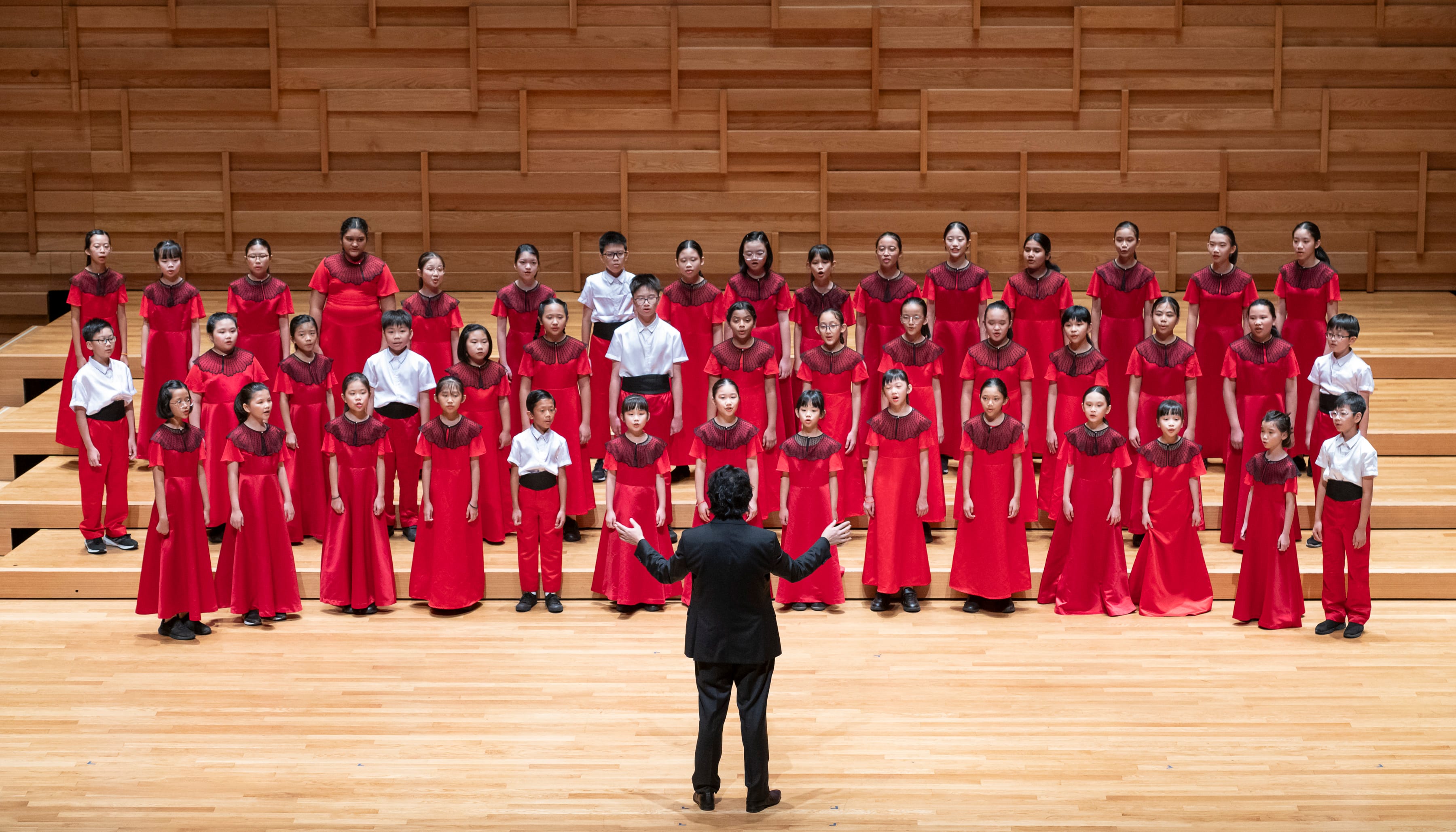 A conductor in black faces a large children's choir in red dresses and white shirts on a wooden stage.
