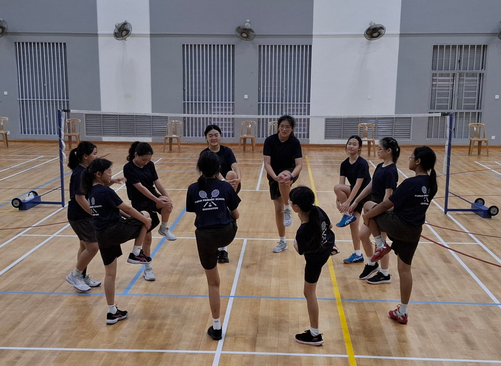A group of young girls in black badminton uniforms stretching in a circle on an indoor court with a net behind them.