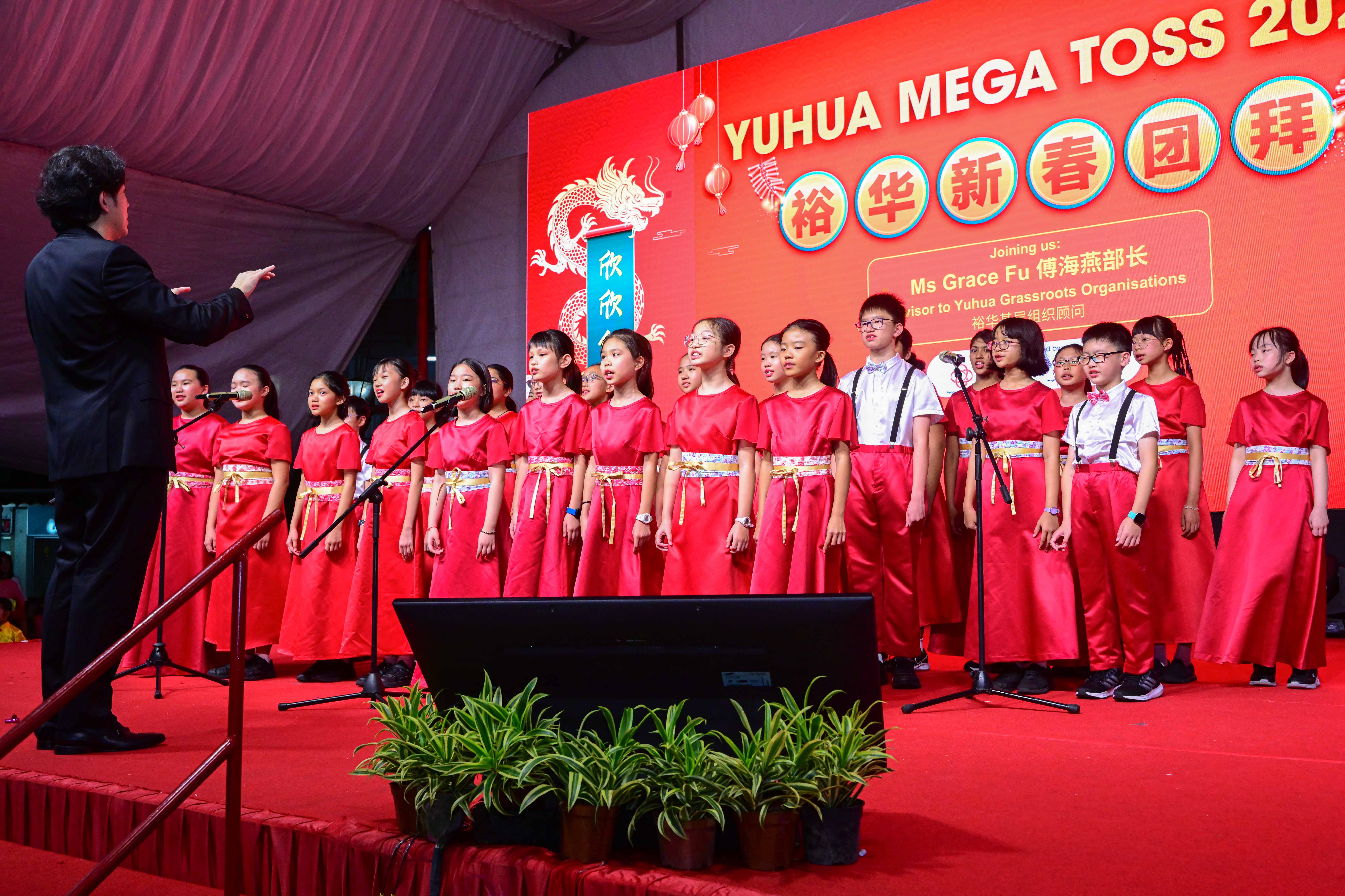 Conductor leads a youth choir in red outfits on stage before a red Yuhua Mega Toss backdrop.