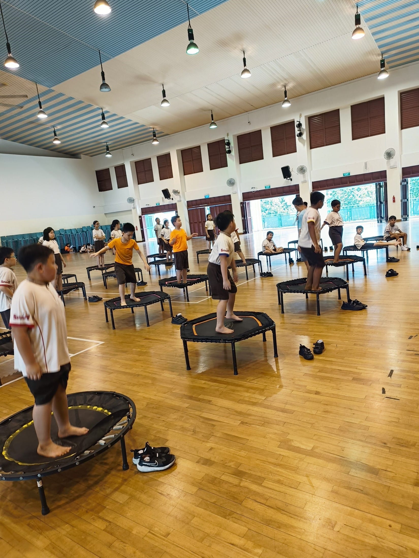 Children in school uniforms stand barefoot on small trampolines spread across an indoor gymnasium floor.