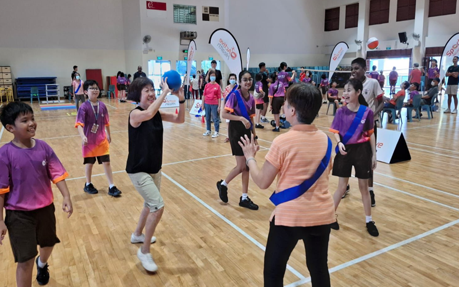 Children and adults play a ball game on an indoor wooden sports court during an organized event.