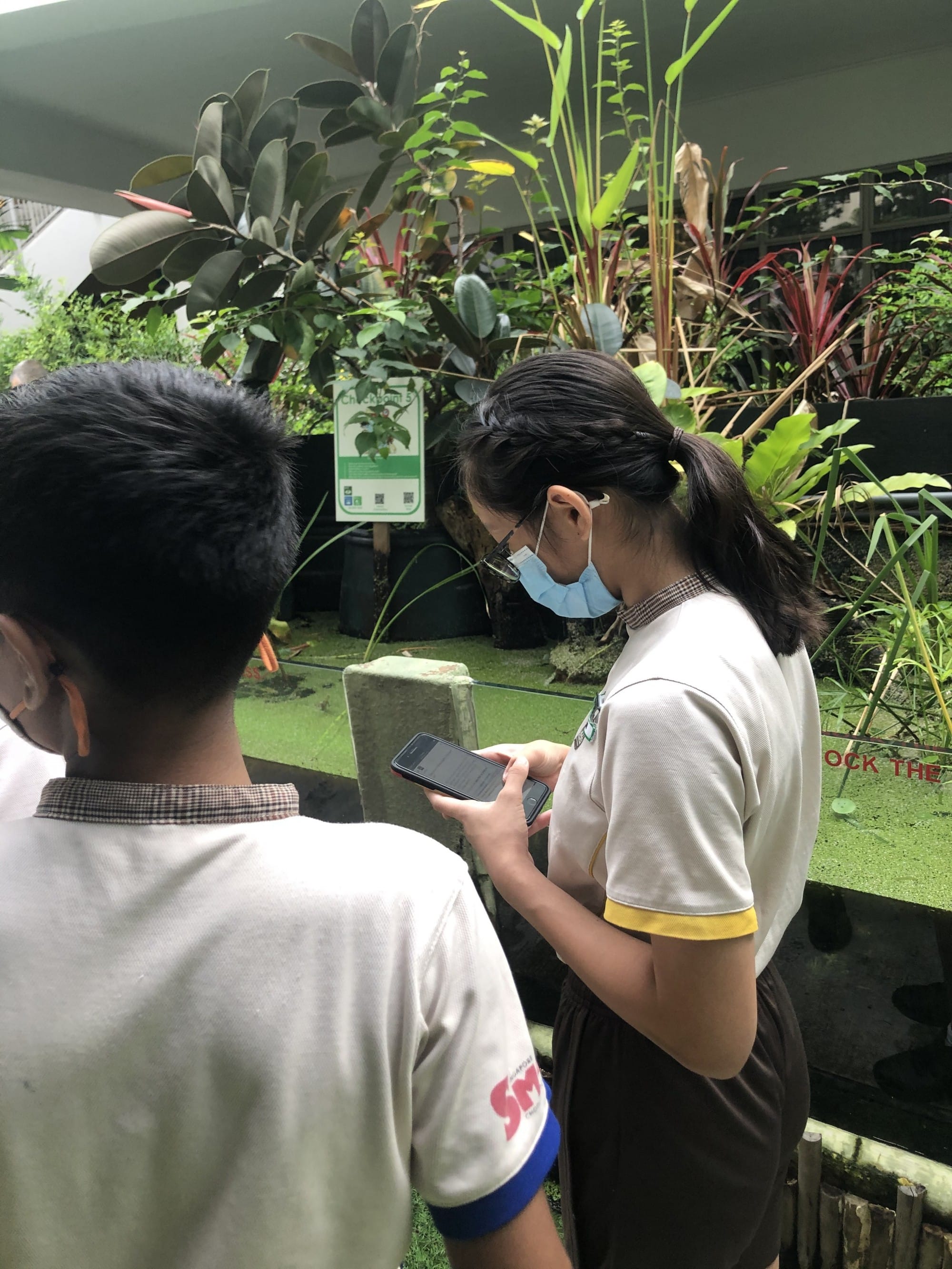 Two masked students in white uniforms look at a phone near a lush garden with tropical plants and a pond.