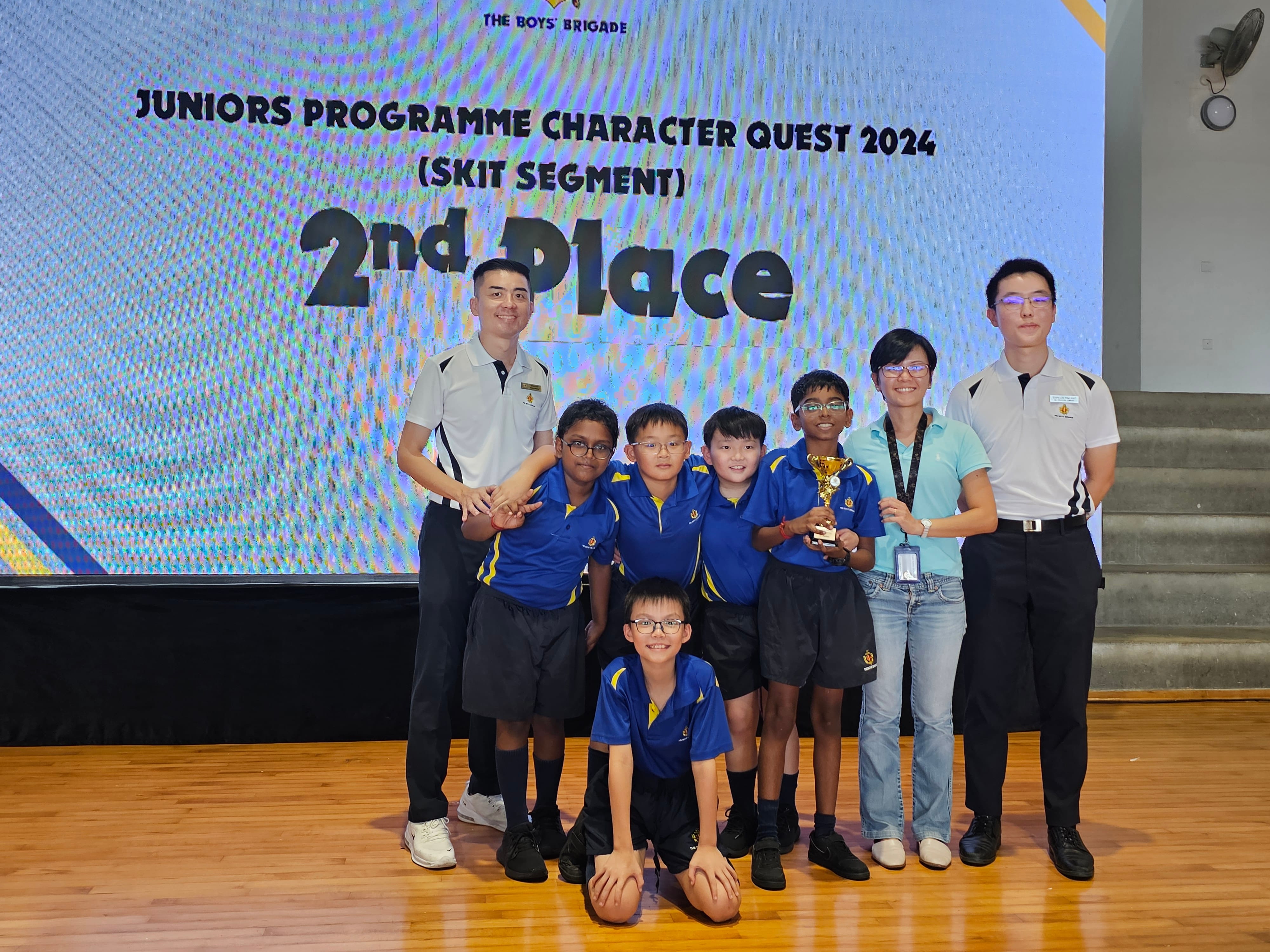 Group of boys in blue uniforms holding a trophy pose in front of a 2nd Place Boys Brigade Character Quest 2024 banner.