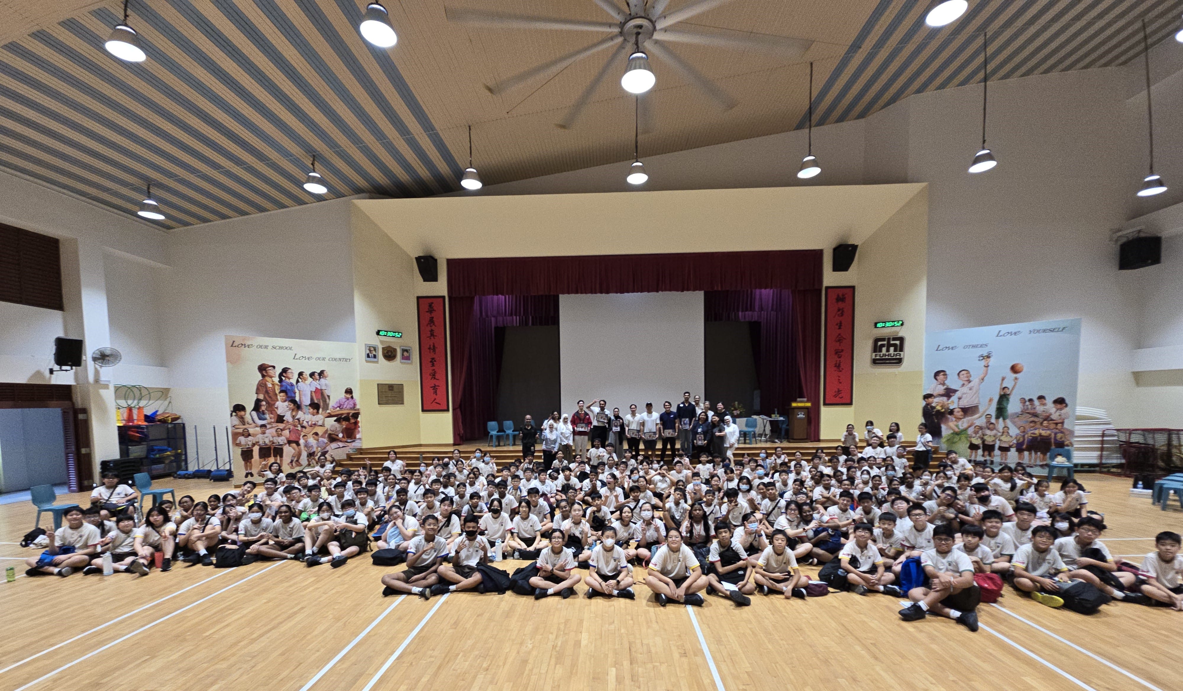 Large group of students in white uniforms sitting on a school hall floor with teachers standing near a stage.