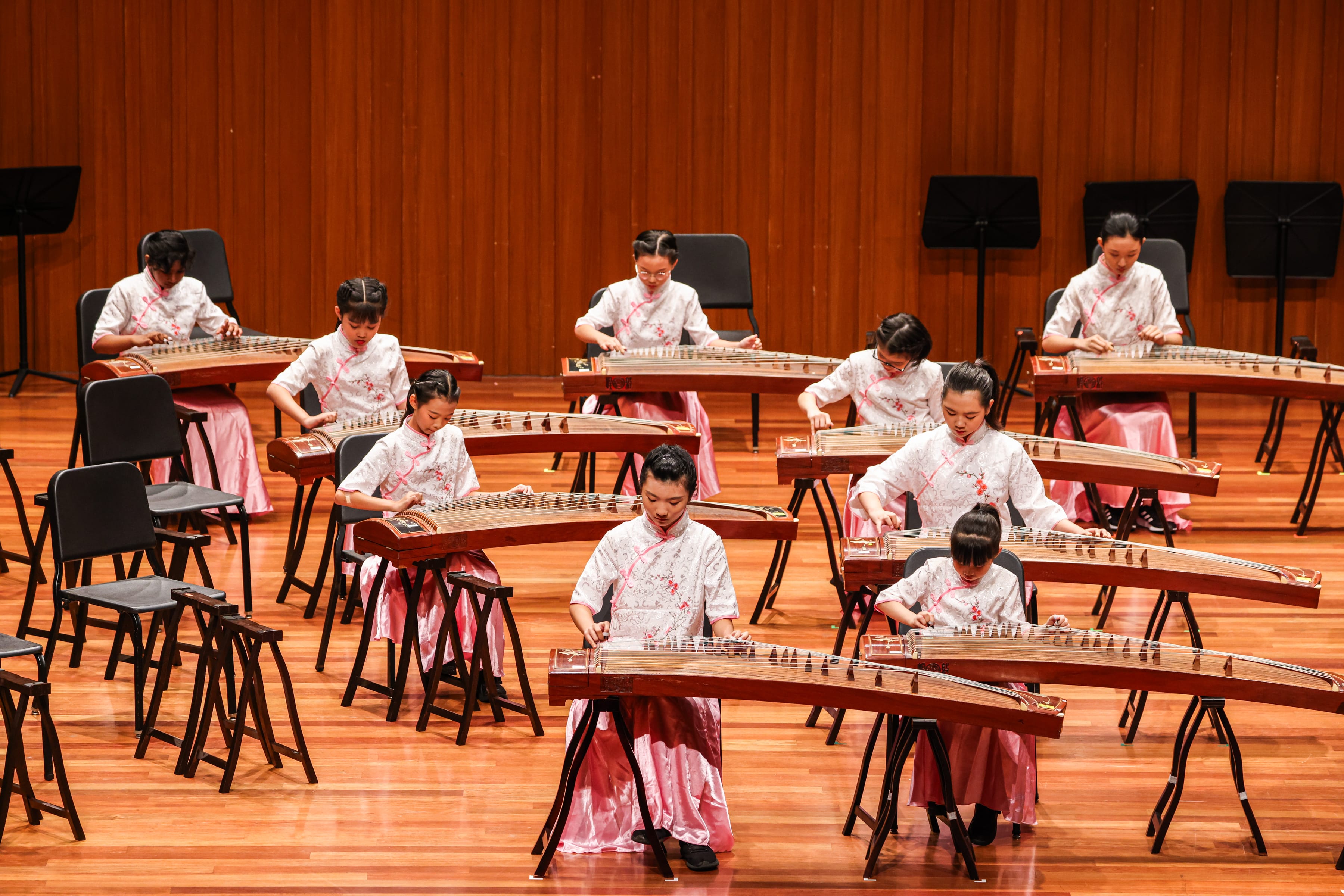 Group of young performers in white and pink traditional outfits playing guzheng instruments on a wooden stage.