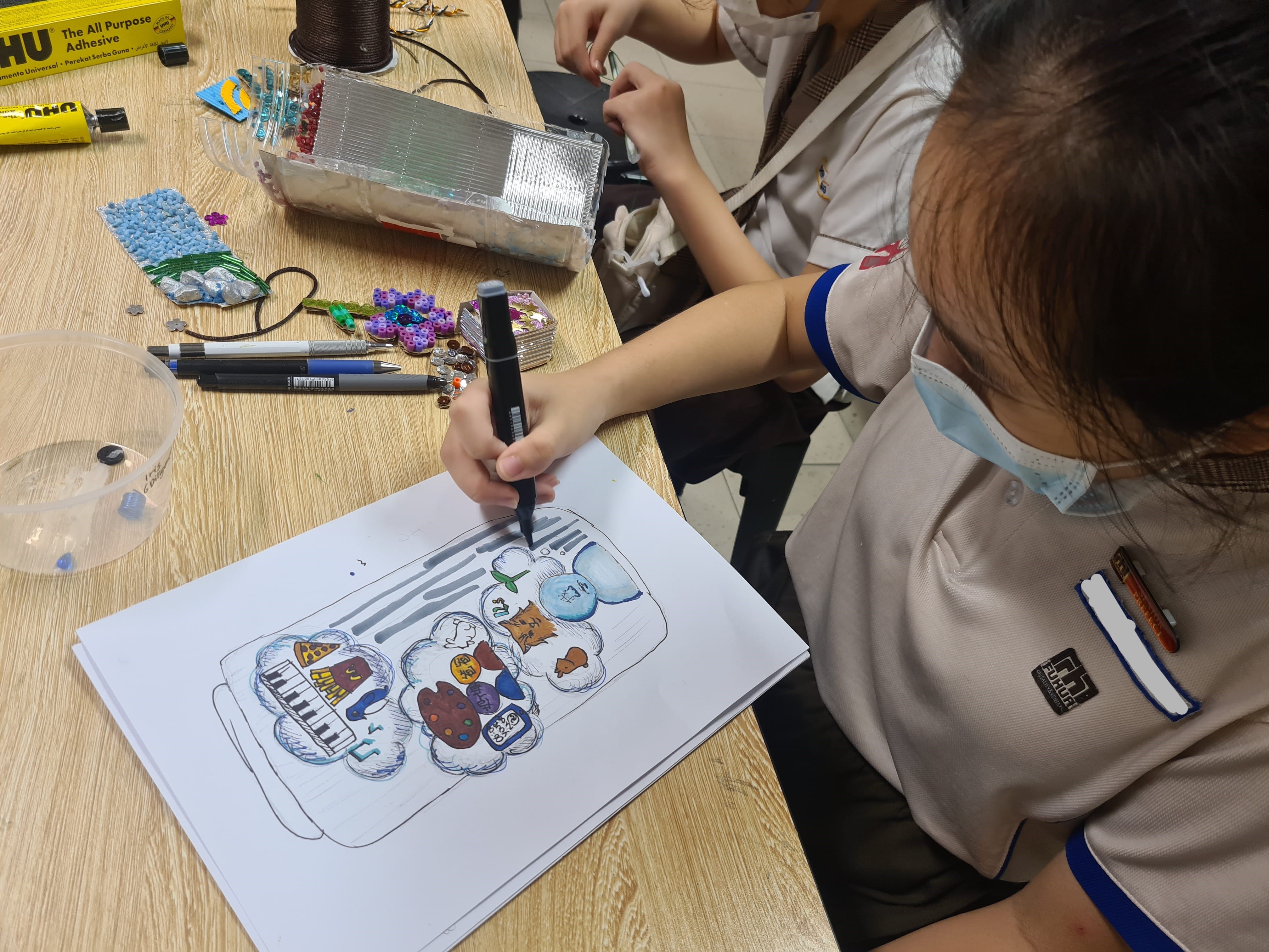 A student in uniform draws a colorful jar illustration with a marker at a craft-filled wooden table.