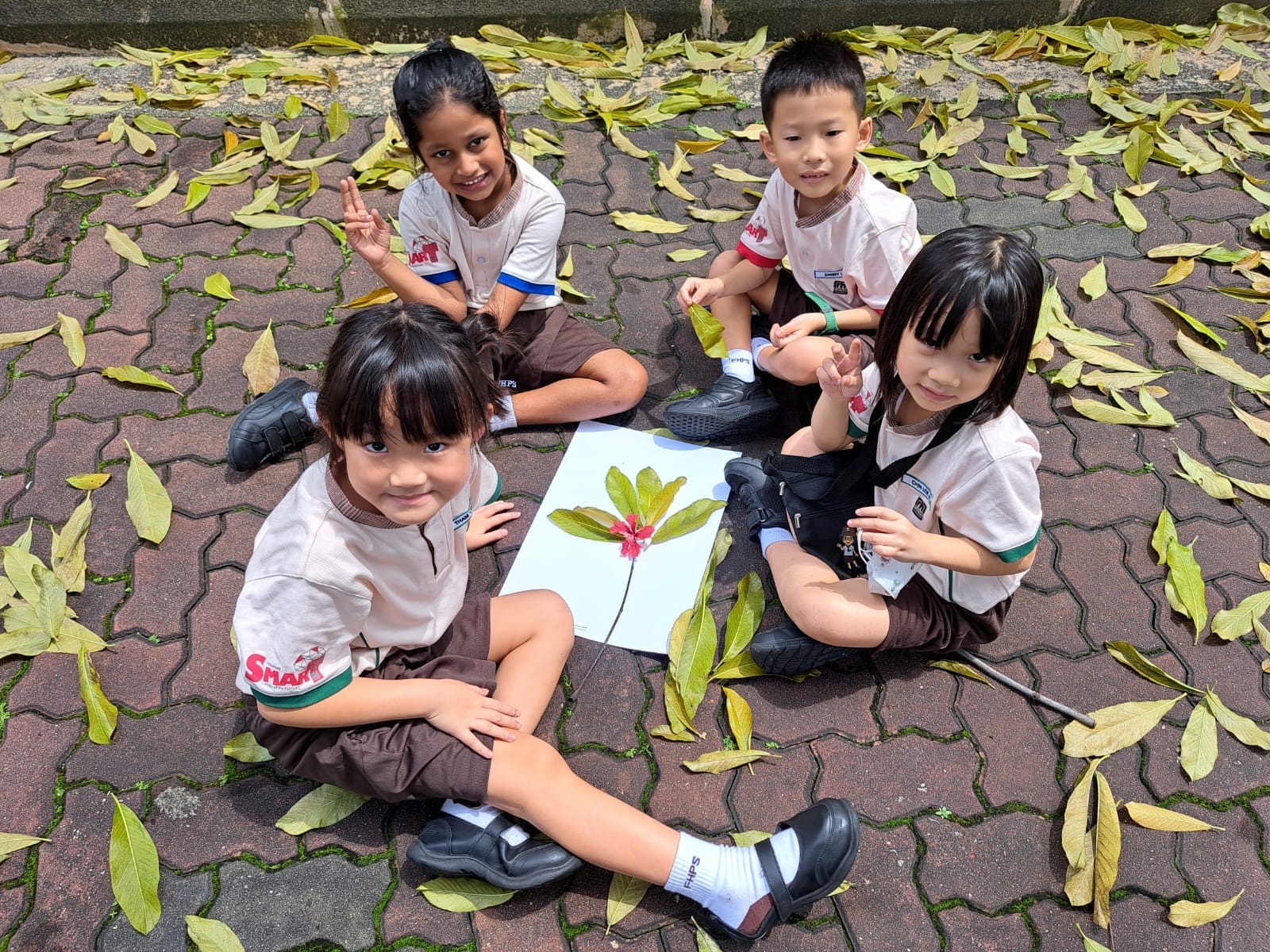 Four young children in school uniforms sit on a brick path covered in fallen leaves, with a leaf and flower arrangement on paper.