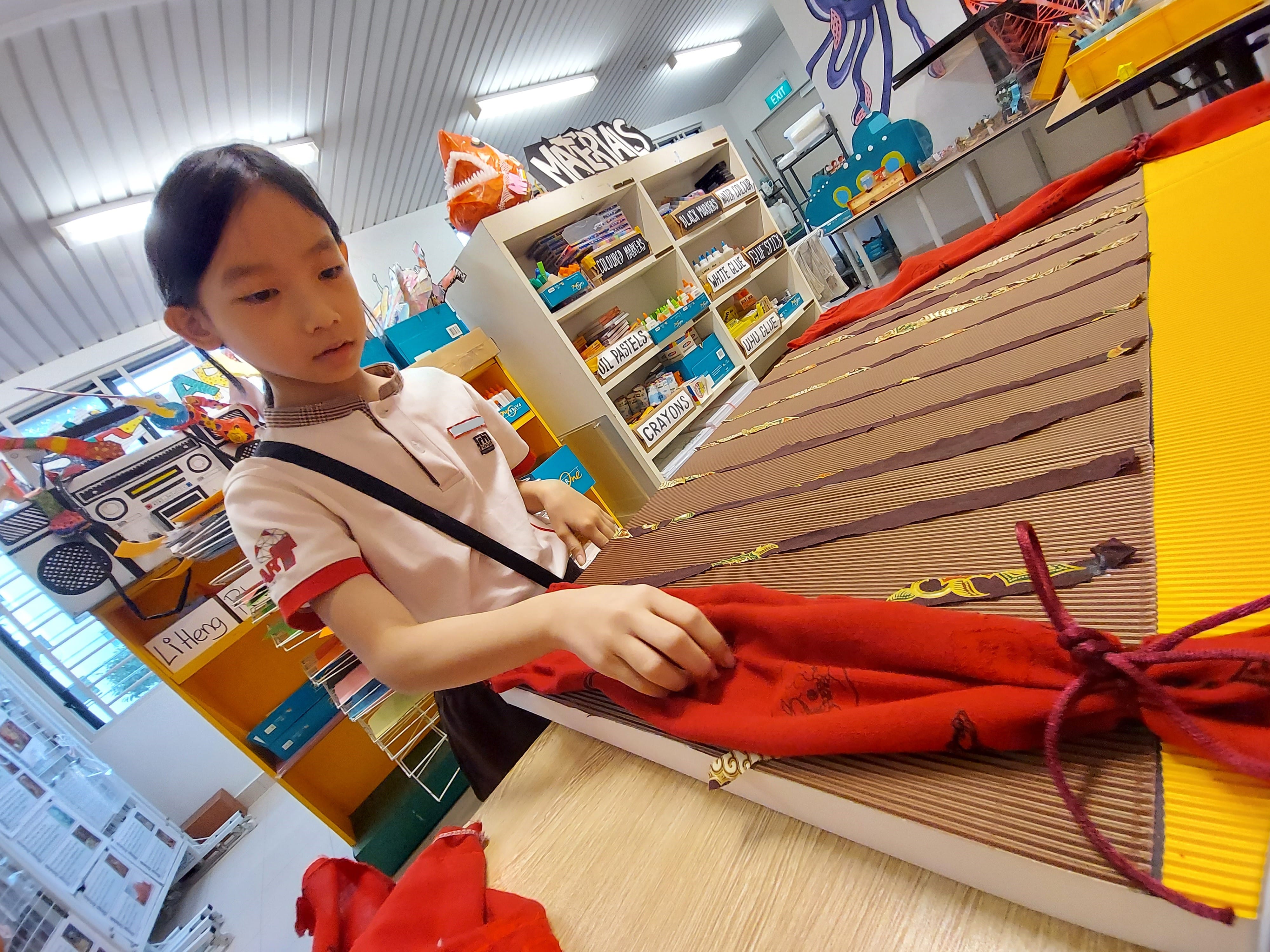 A young girl in school uniform handles red fabric on a cardboard craft project in a colorful art classroom.