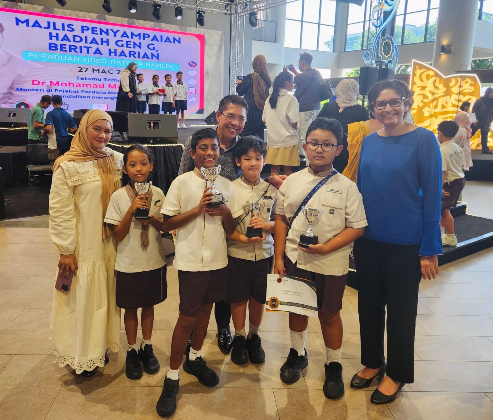 Four uniformed children hold trophies with two adults at a Majlis Penyampaian Hadiah Gen G award ceremony.
