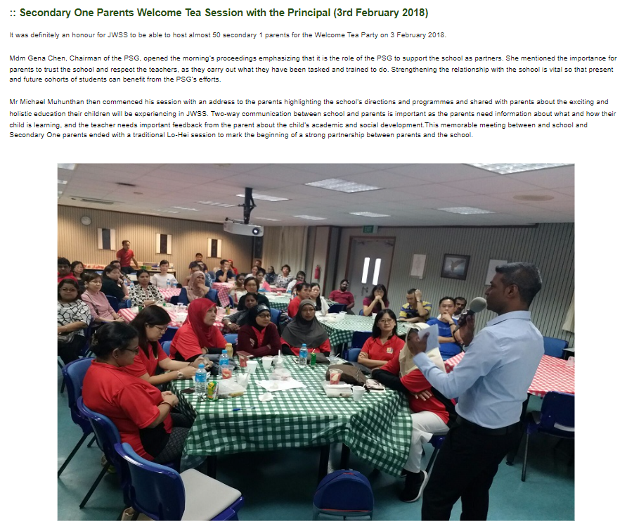 A man speaks to a large group of seated adults at checkered tables in an indoor hall.