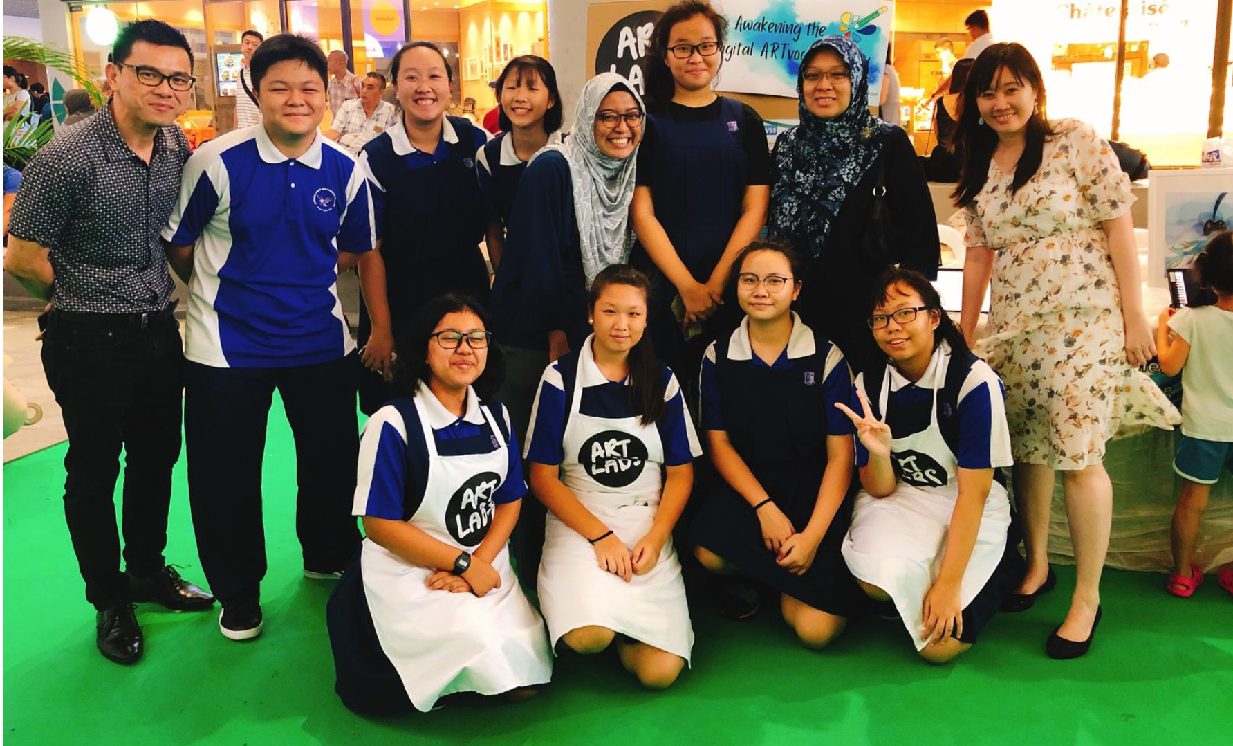 Group of 11 people posing indoors; several in blue uniforms and white aprons on a green floor.