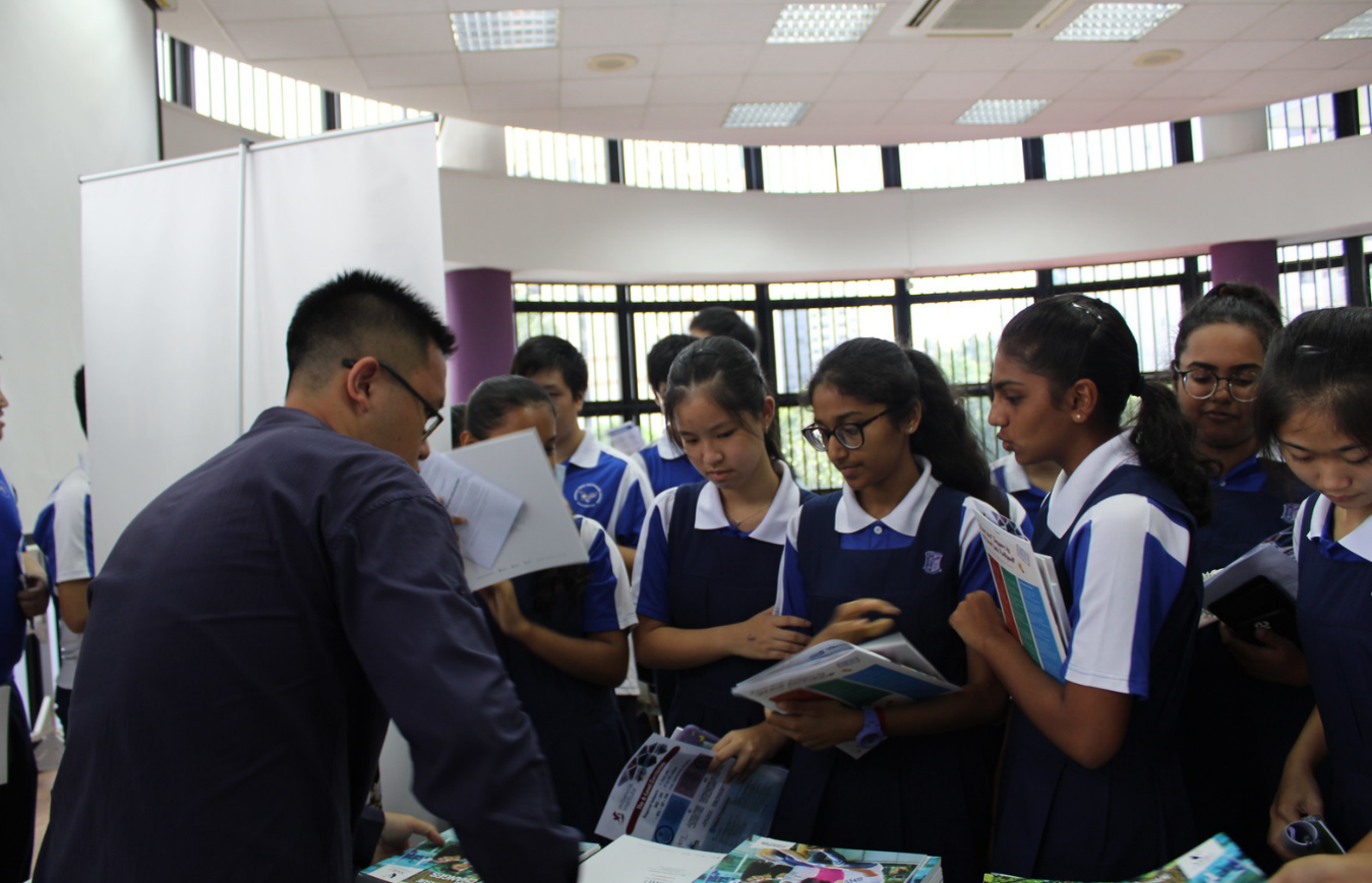 Students in blue and white uniforms gather around an adult reviewing printed brochures indoors.