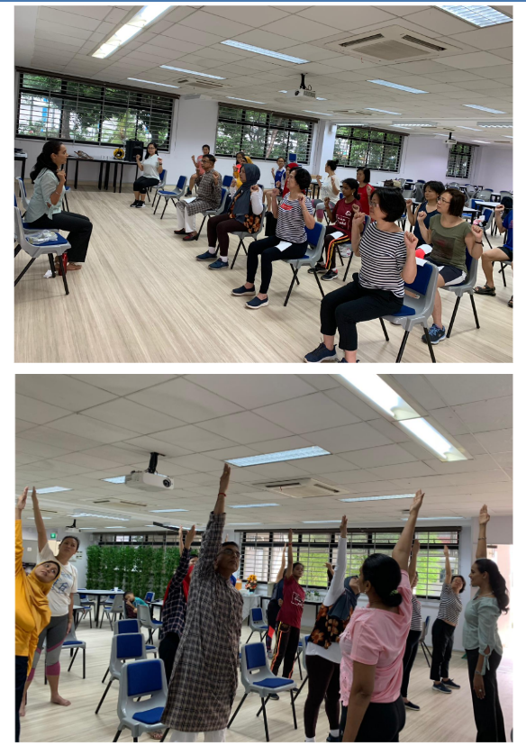 Group of people doing seated and standing exercises in a bright indoor room with chairs arranged in rows.