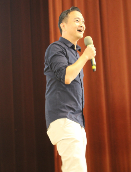 Man in navy shirt and white pants smiling while holding a microphone on stage with a red curtain behind him.
