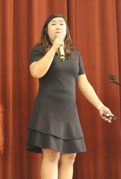 Woman in a black dress holding a microphone and speaking on stage in front of a red curtain.