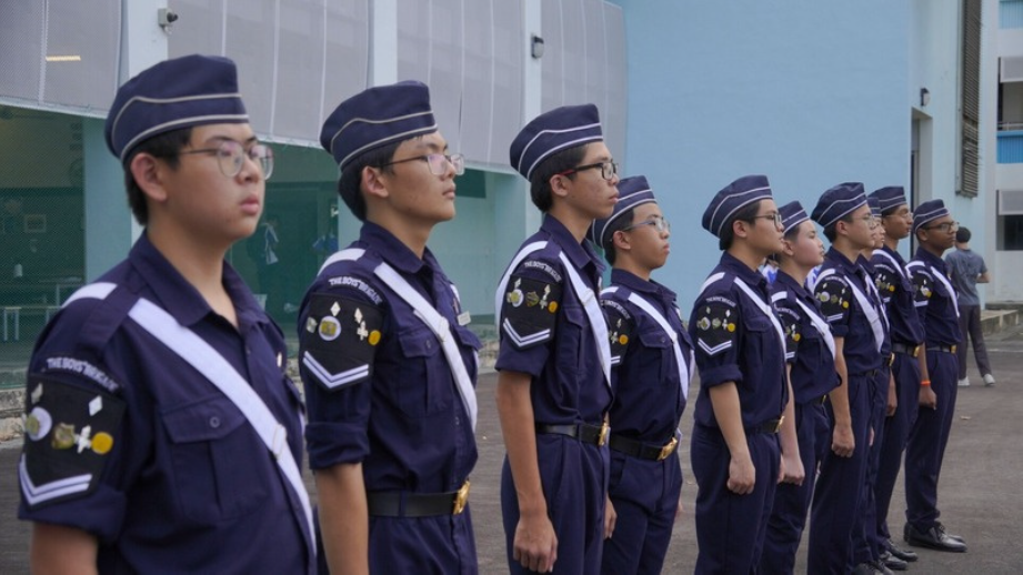 A row of uniformed cadets in navy blue stand at attention outdoors beside a building.
