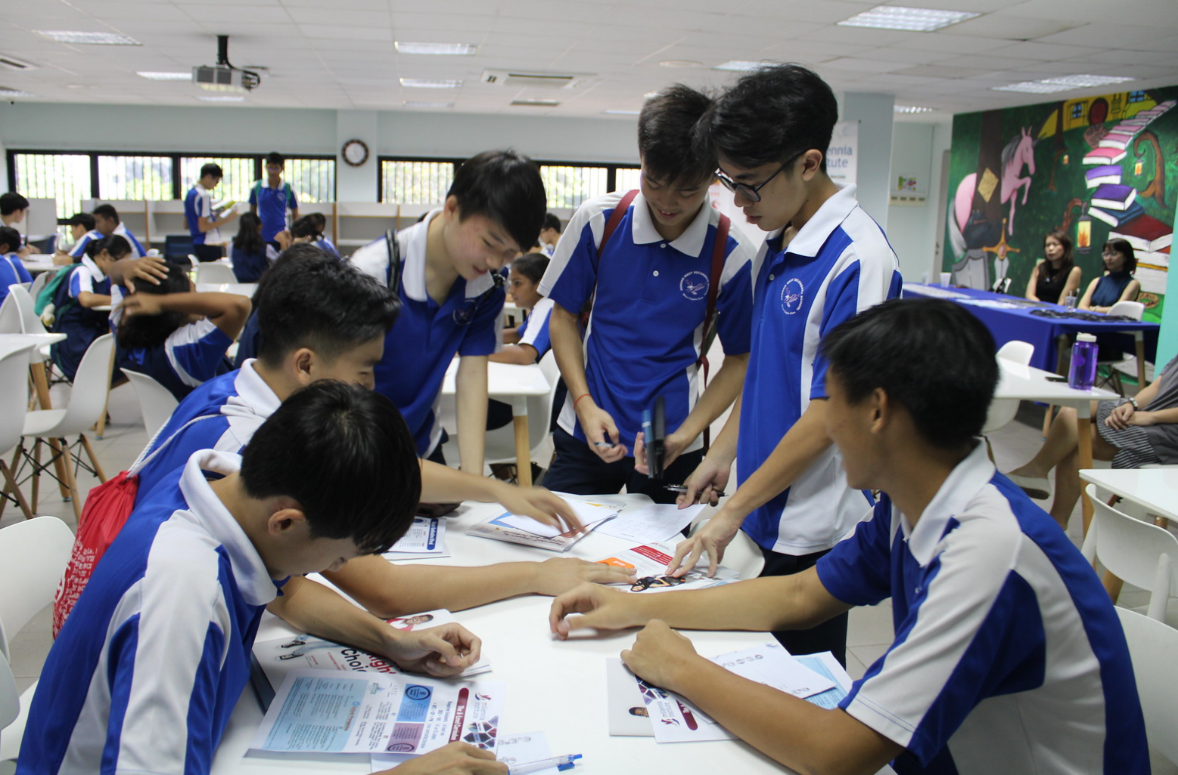 Several students in blue and white uniforms gather around a white table, reviewing printed papers and brochures together.