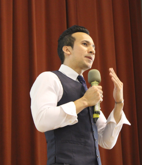 A man in a white shirt and navy vest speaks into a microphone in front of a red curtain.
