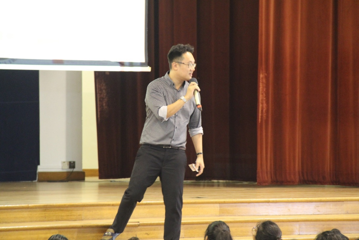 A man wearing glasses and a gray shirt holds a microphone while speaking on a wooden stage with a projection screen behind him.