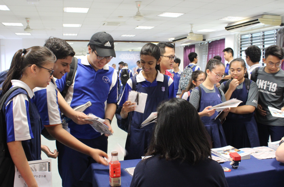Students in blue uniforms gather around a table at an indoor fair, holding brochures and talking to a seated person.