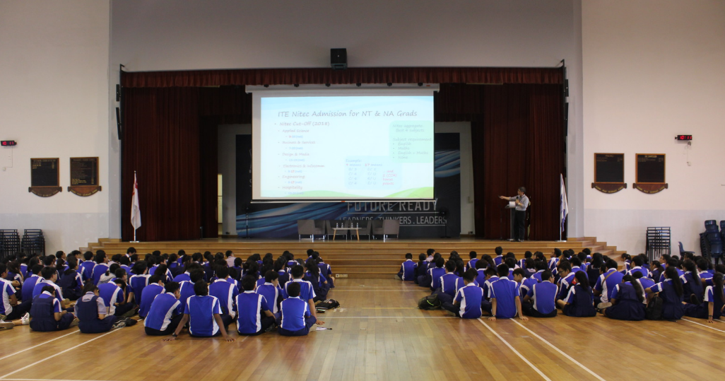 Students in blue uniforms sit on a gymnasium floor watching a presenter at a stage with a projected slideshow.