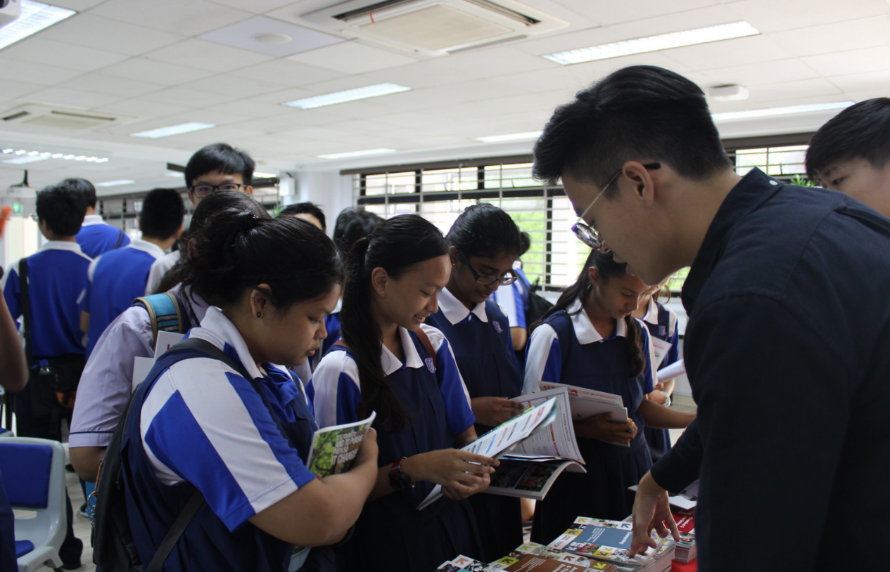 Students in blue uniforms browse brochures at an indoor fair while an adult in black points to materials on a table.