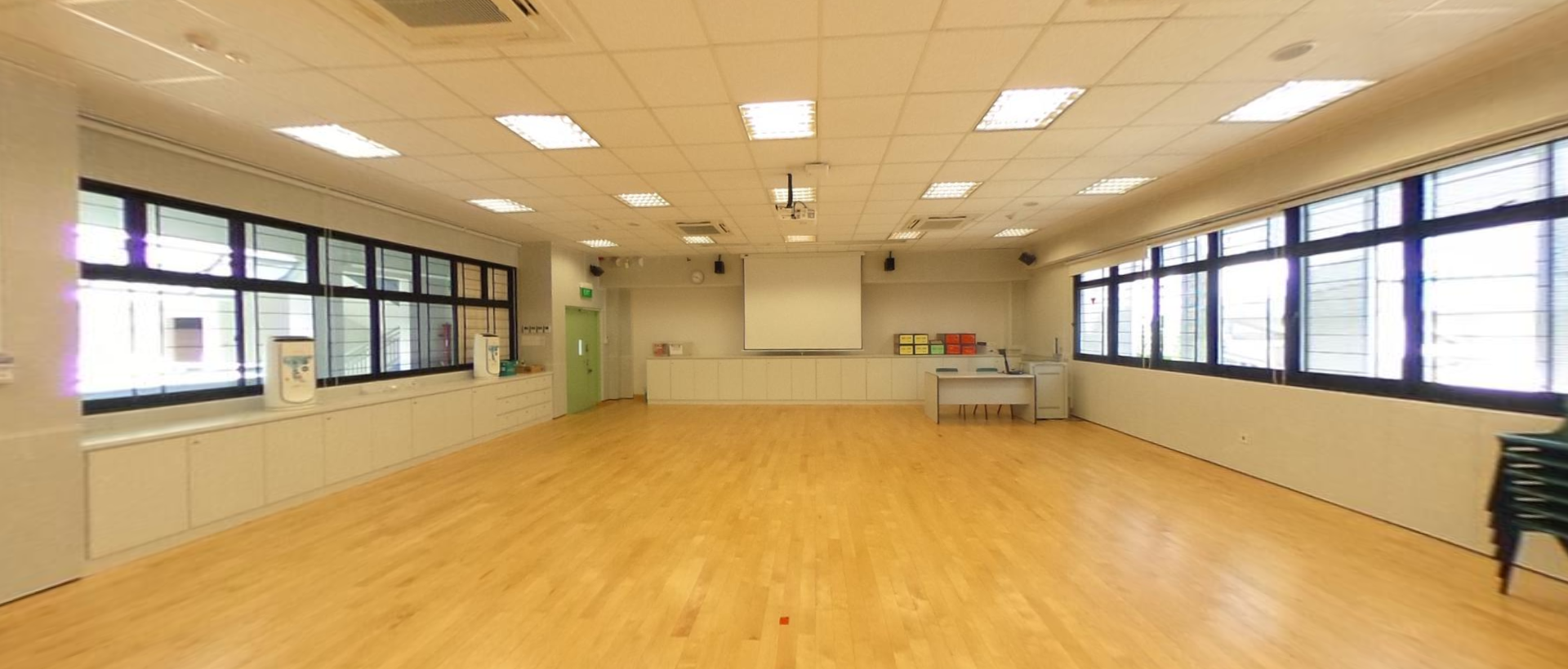 Empty room with wood floor, white ceiling tiles, fluorescent lights, a projector screen, and windows along both walls.