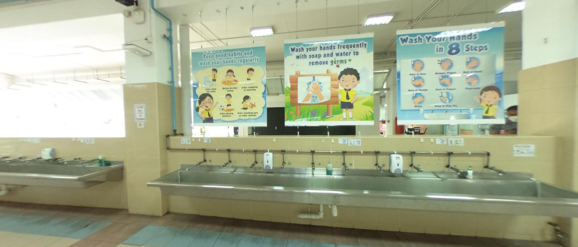 A row of stainless steel sinks with three handwashing instruction posters mounted on the wall above them.