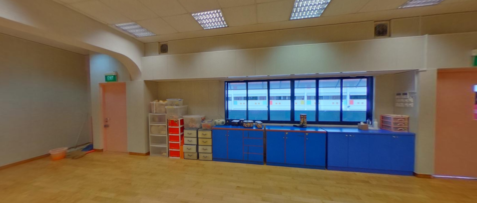 Spacious classroom with blue storage cabinets, plastic drawer units, and a wide window along the back wall.