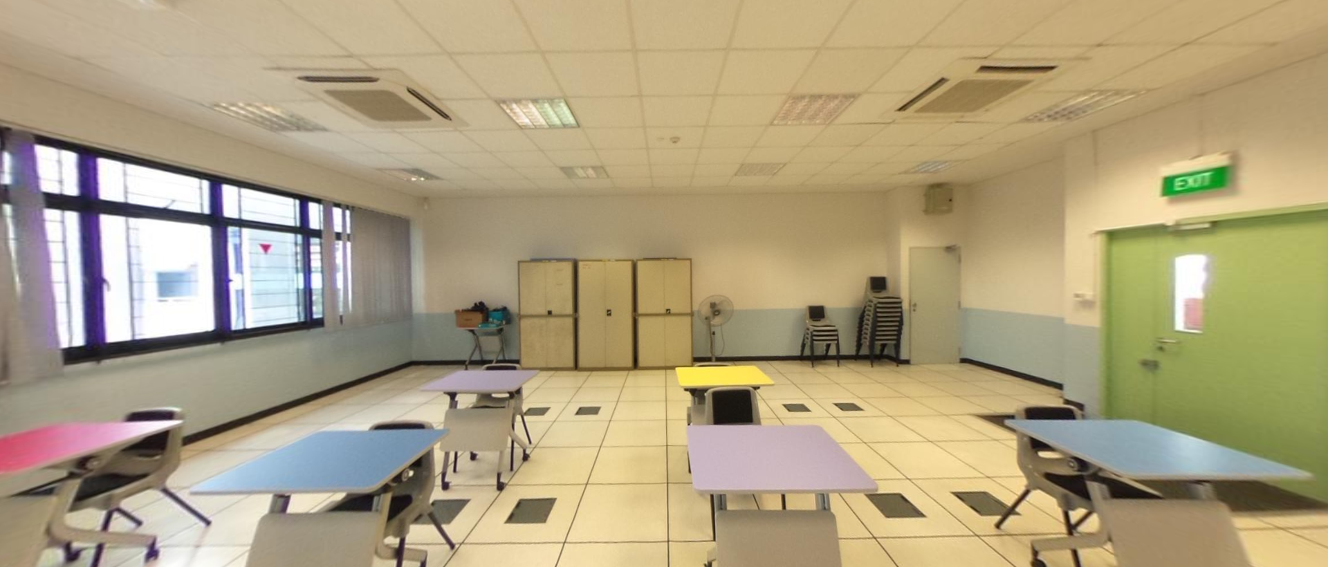 Empty classroom with colorful tables in pink, blue, yellow, and purple, chairs, cabinets, and a green exit door.