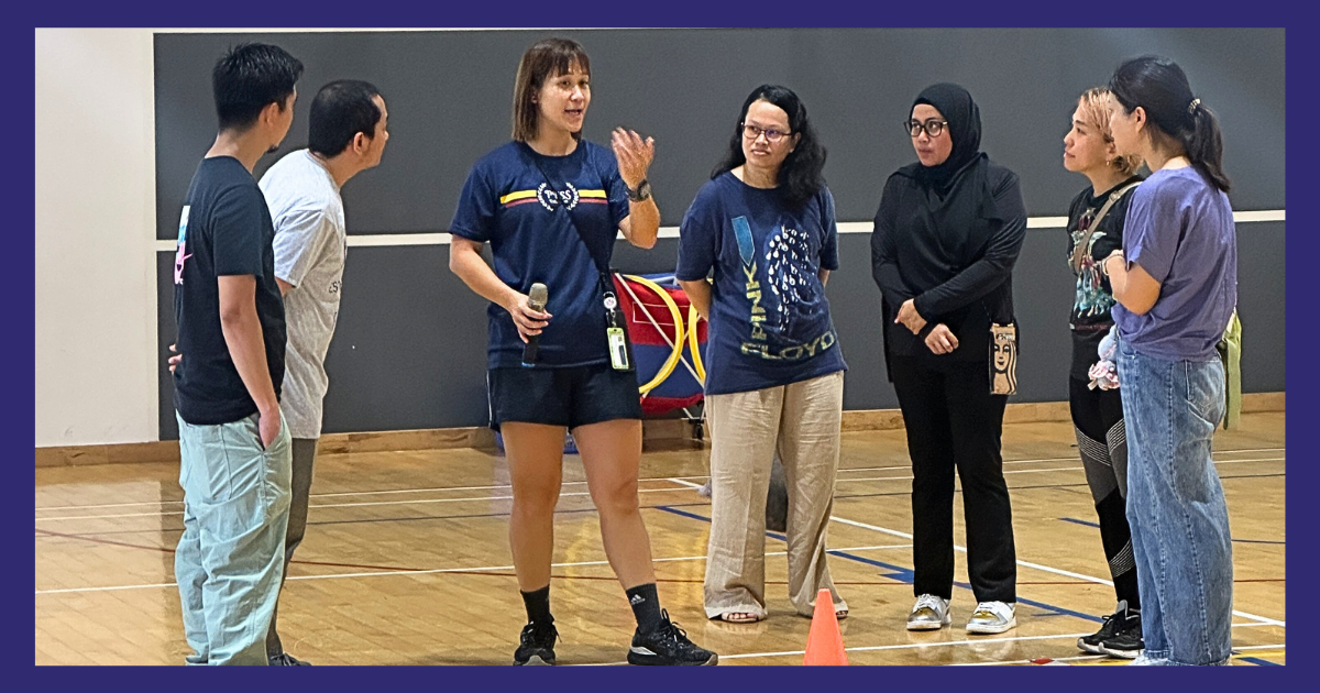Six people stand in a circle on a wooden gym floor, with one woman holding a microphone and speaking.