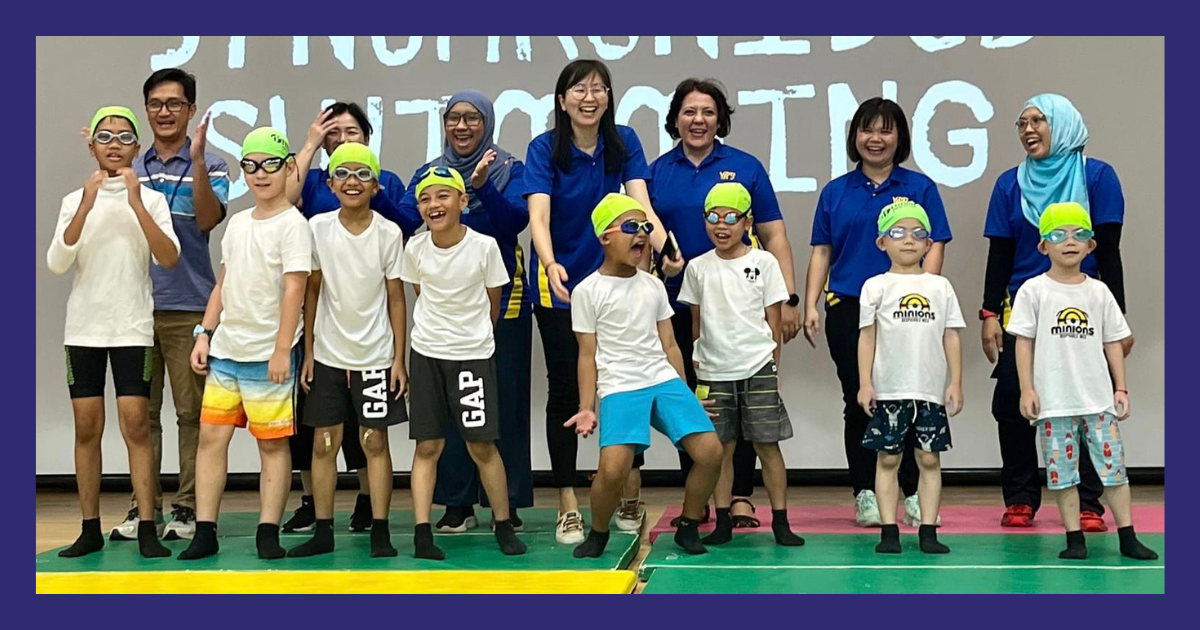 Group of children wearing swim goggles and caps posing with adults in blue shirts before a synchronized swimming banner.