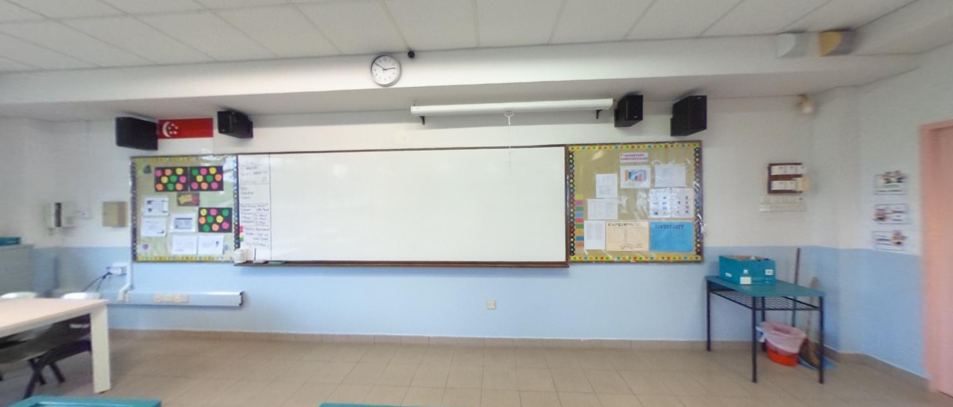 Empty classroom with a large whiteboard, bulletin boards, a wall clock, speakers, and a small shelving unit.