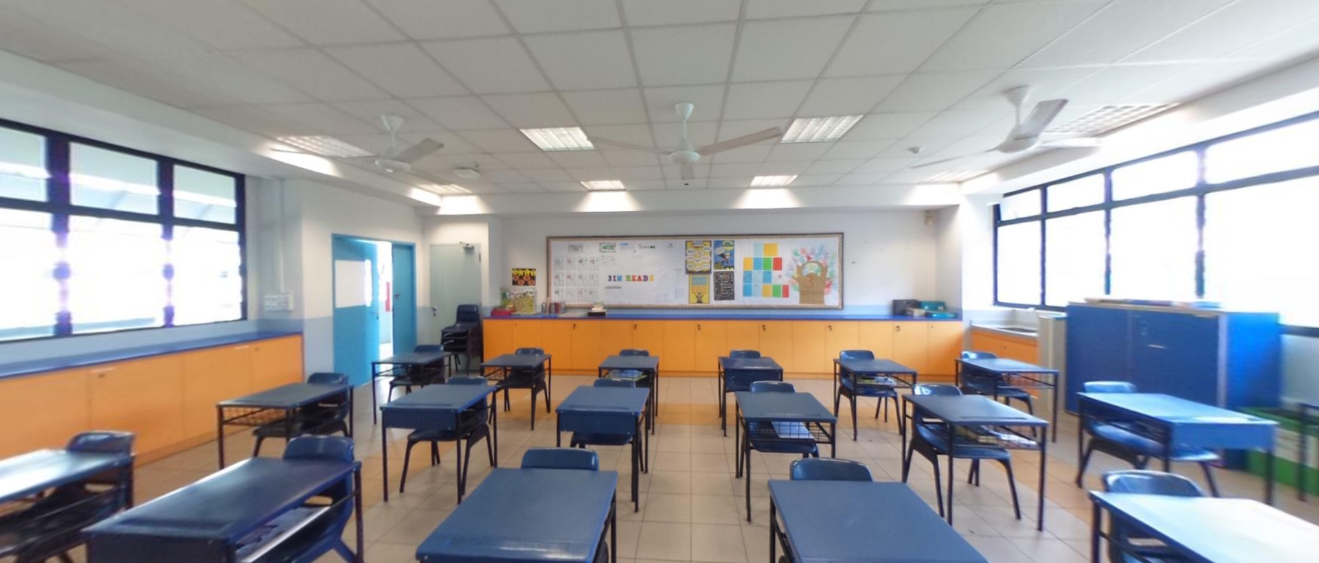 Empty classroom with blue desks and chairs, orange cabinets, a bulletin board, ceiling fans, and large windows.