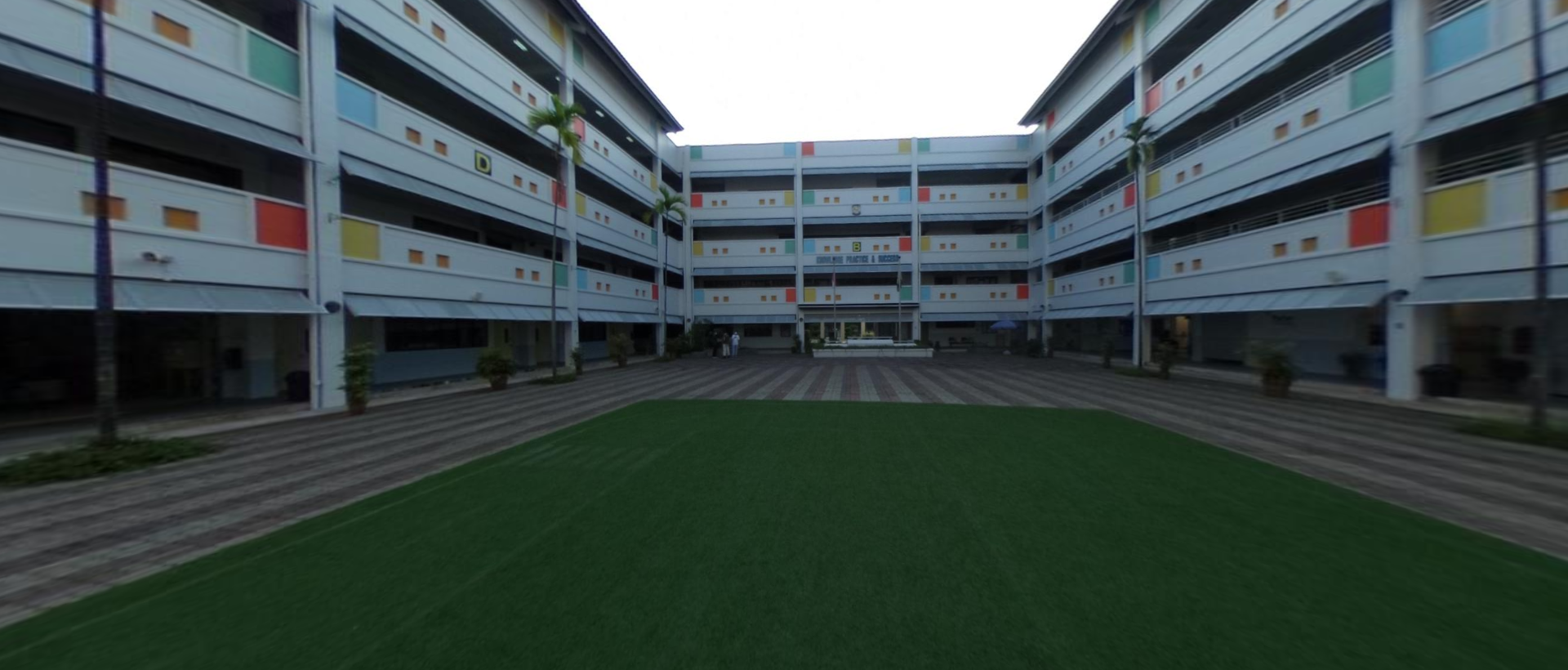 A multi-story school building with colorful panels forms a U-shape around a green courtyard with paved pathways.