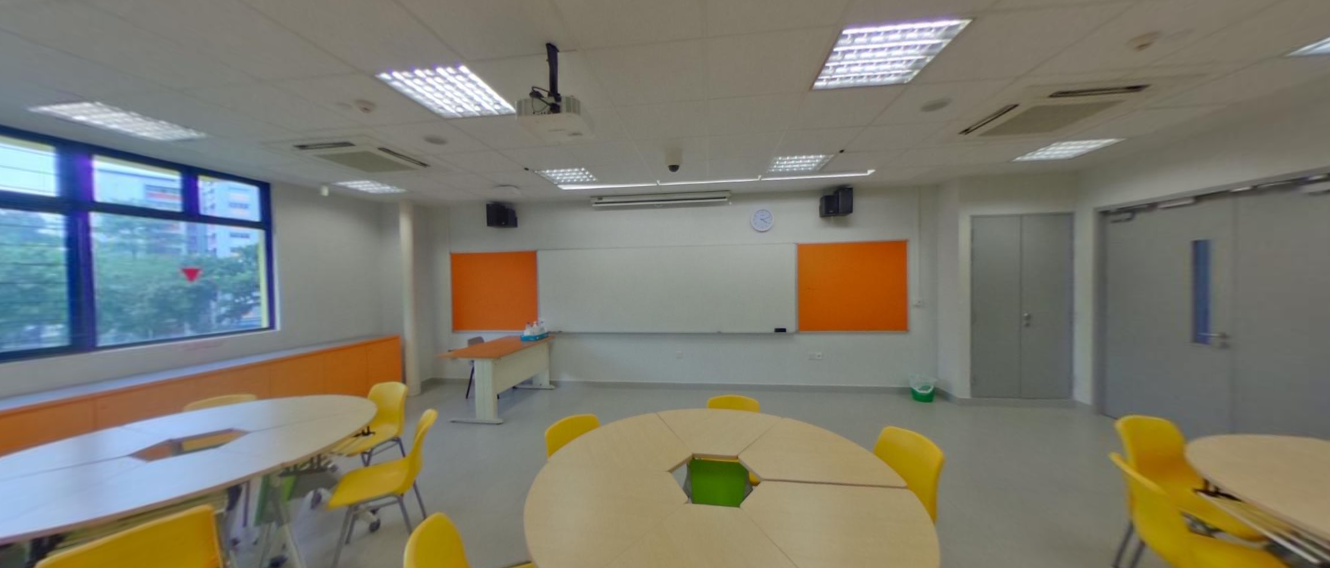 Empty classroom with round tables, yellow chairs, orange wall panels, a whiteboard, and a ceiling-mounted projector.