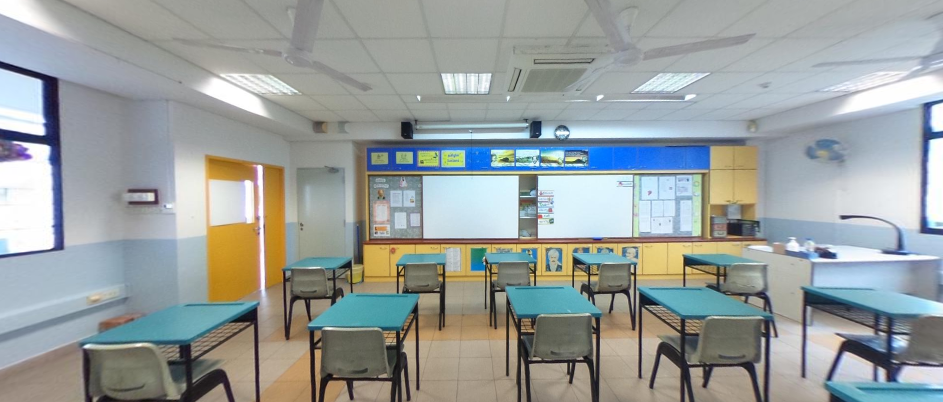 Empty classroom with teal desks, gray chairs, a whiteboard, yellow cabinets, and a blue display board at the front.