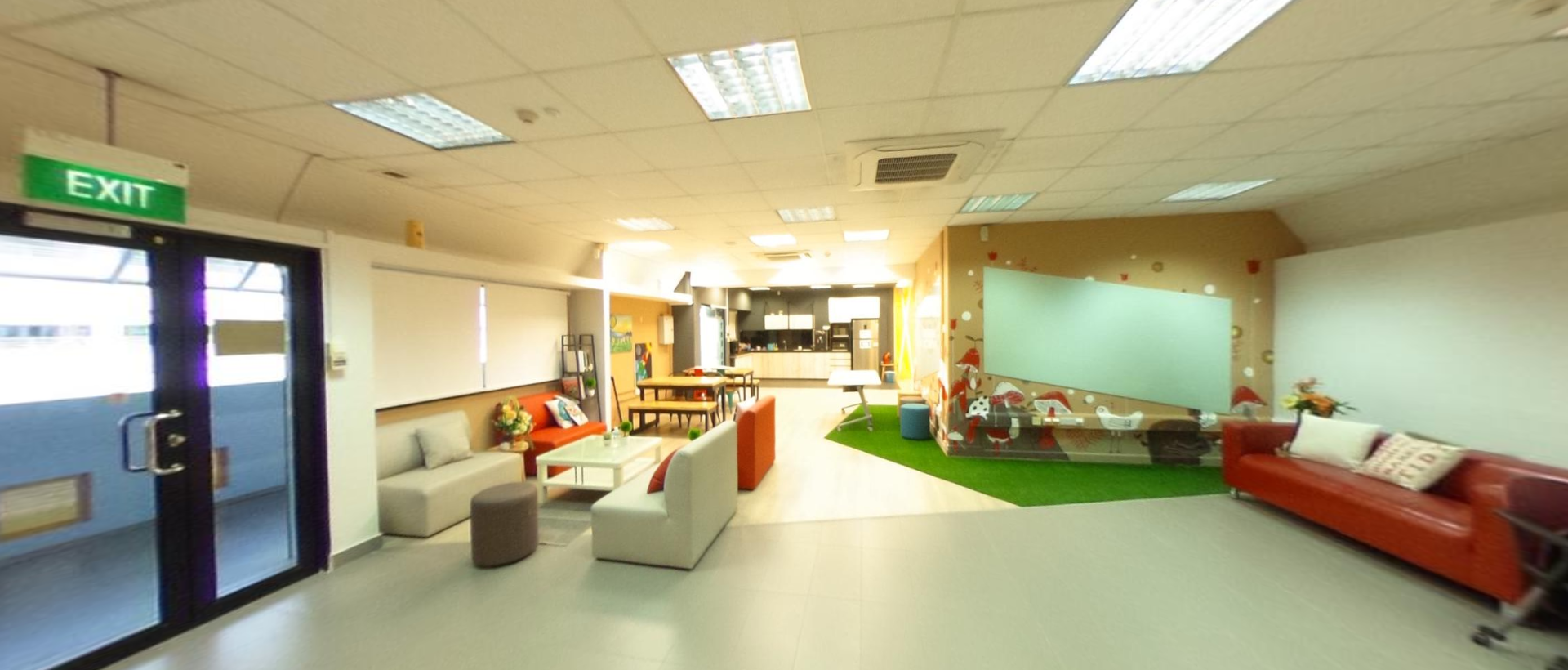 Bright open common area with gray and red sofas, a green turf patch, whiteboard, and a kitchenette in the background.