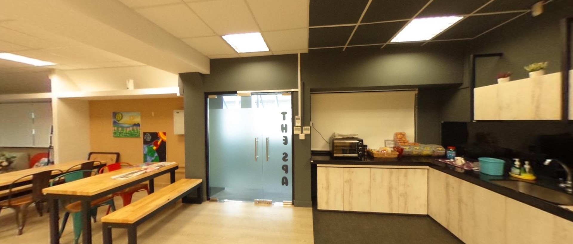 Office break room with wooden tables, benches, a kitchen counter with a toaster oven, and frosted glass doors.