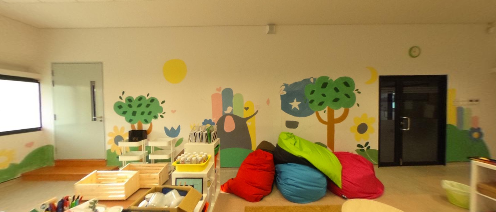 Bright children's classroom with colorful bean bags, wooden crates, shelves, and a nature-themed mural on the wall.