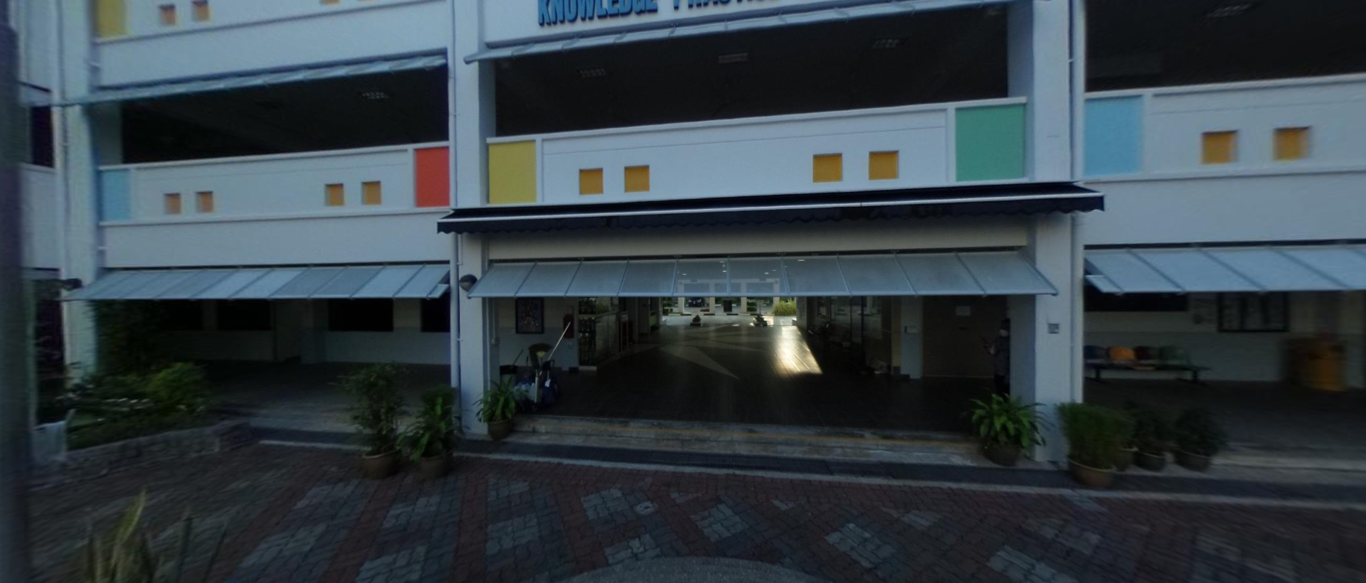 Multi-story building entrance with awnings, colorful panels, potted plants, and a brick-paved courtyard.