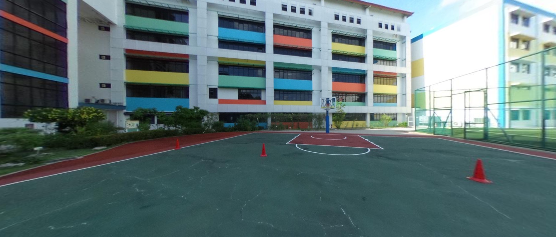 Outdoor basketball court with orange cones in front of a colorful multi-story school building.