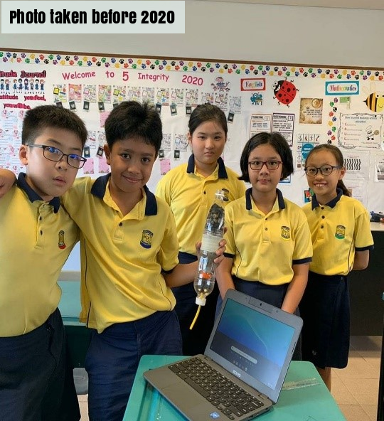 Five students in yellow uniforms pose in a classroom with a laptop and a bottle device on a desk.