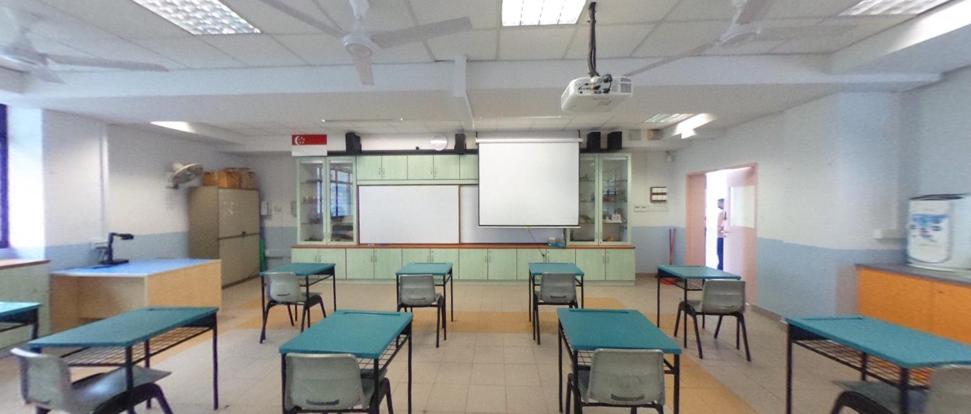 Empty school science classroom with teal desks, chairs, a whiteboard, projector screen, and green storage cabinets at the front.