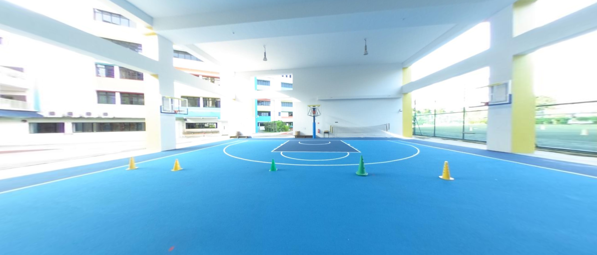 Covered blue basketball court with yellow and green training cones, a hoop, and surrounding school buildings.