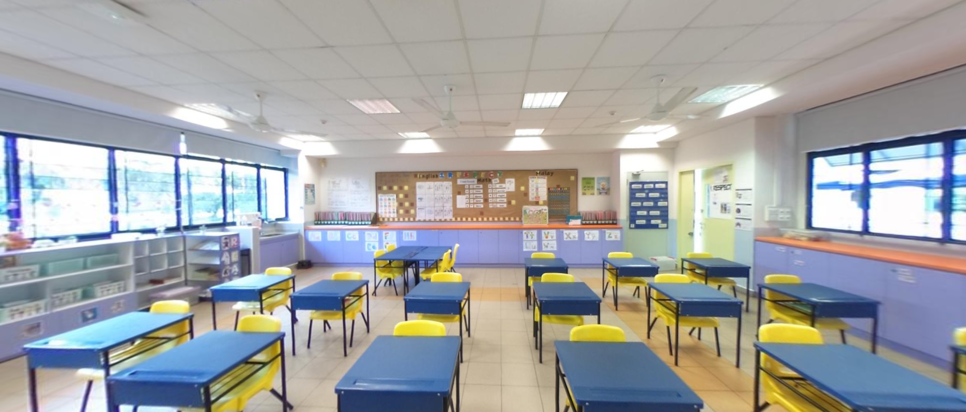 Empty classroom with rows of blue desks and yellow chairs, a bulletin board at the front, and large windows on the left.