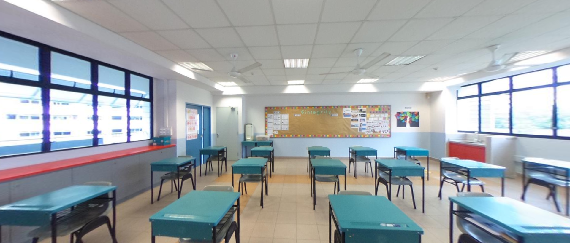 Empty classroom with rows of teal desks and chairs, a bulletin board on the back wall, and large windows on both sides.